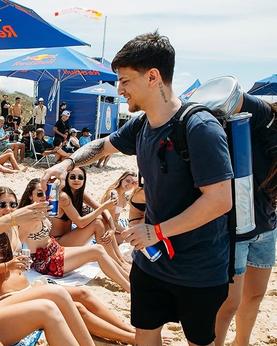 Arthur Lacerda, de camisa azul-marinho, entregando latas a pessoas numa ativação na praia.